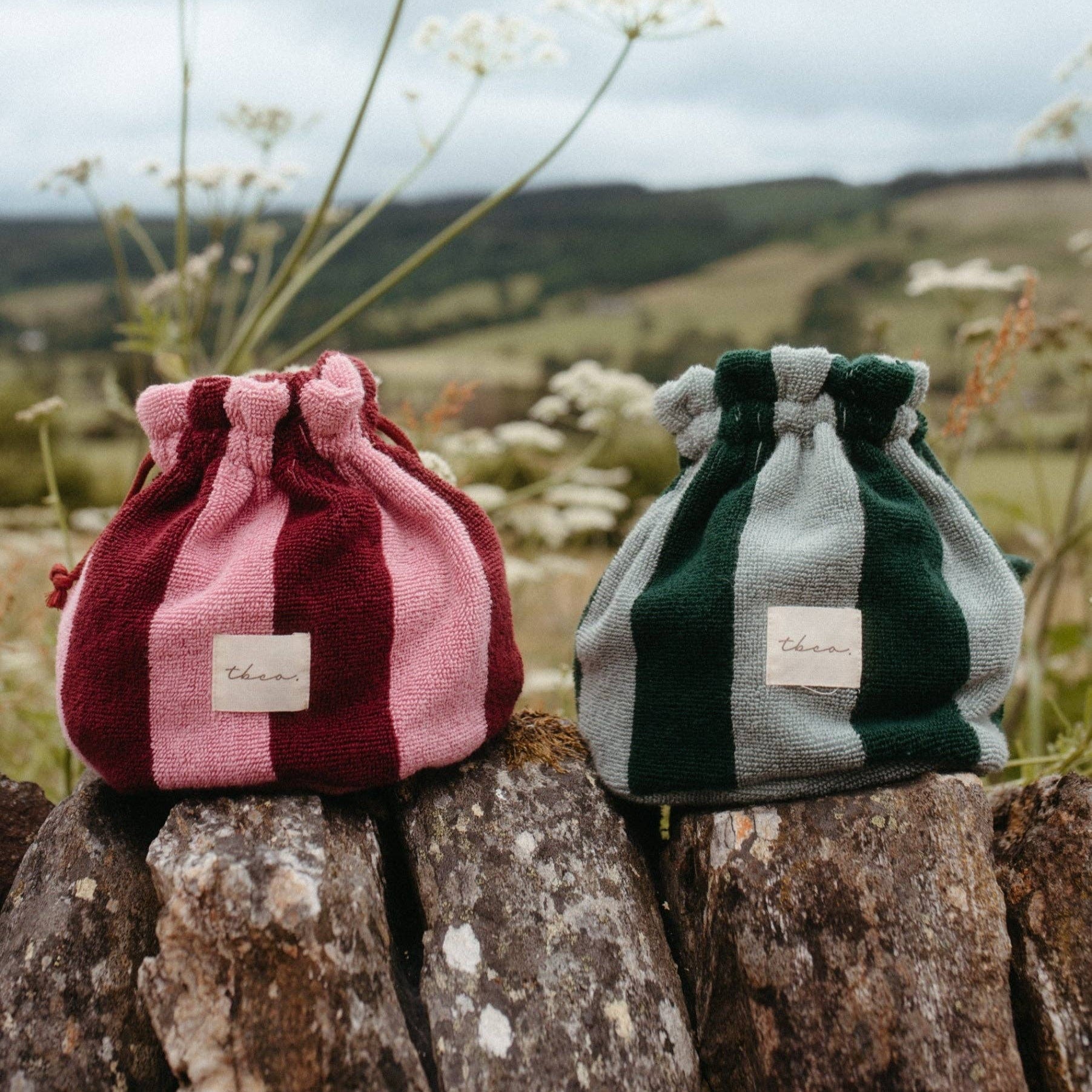 Two drawstring bags, one pink and one green and white checkered, on a stone surface with a scenic background.