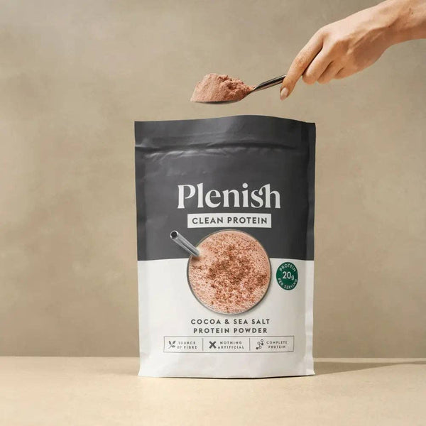 A stand-up pouch of Plenish Clean Protein Cocoa & Sea Salt Protein Powder on a beige surface, with a hand holding a spoon above it containing some of the powder.