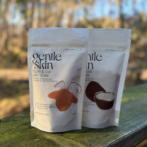 Two stand-up pouches of Gentle Skin CoCoa & Oat Bath Soak standing on a weathered wooden surface outdoors with a blurred forest background.